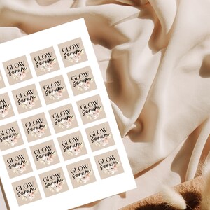 May include: A sheet of square stickers with the words "GLOW Serum" in a handwritten font. Each sticker has a beige background and a floral design. The stickers are arranged on a white sheet, with a soft, neutral-toned fabric in the background.