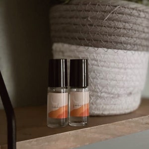 Op de afbeelding: Twee kleine, heldere glazen flesjes met zwarte doppen en etiketten met oranje en beige ontwerpen. De flesjes staan op een houten plank, met een gevlochten mand op de achtergrond. De etiketten lezen "calm" en "focus".