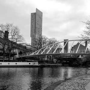 May include: Black and white photograph of a city canal scene. A white pedestrian bridge arches over the water, with a tall modern building in the background. A boat is docked on the canal.
