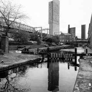 May include: Black and white photograph of a canal scene with a modern skyscraper reflected in the water. A bridge and older brick buildings are also visible, with a cloudy sky overhead. The image is a study in contrasts.