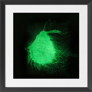 May include: A vibrant image captures the moment a green water balloon bursts, with water droplets radiating outwards against a black background. The photograph is framed in a black square.