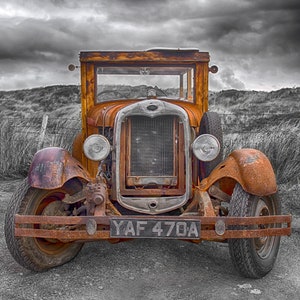 May include: A vintage, rusty orange truck with a wooden cab and a weathered appearance. The front features a prominent grill, headlights, and a license plate. The background is a grayscale landscape with a cloudy sky.