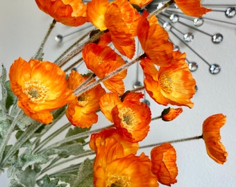 May include: Close-up of vibrant orange poppy flowers with textured petals and yellow centers. The flowers are on slender stems with silvery-green leaves. The background features a decorative silver burst.