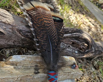 May include: A handcrafted feather wand with dark brown and tan patterned feathers. The handle is wrapped with red and blue cord, and decorated with beads and a leather cord. The wand rests on driftwood and a white fur-like surface. A decorative item.