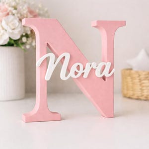 Peut inclure: Une lettre "N" en bois rose avec le nom "Nora" en écriture blanche. La lettre est debout sur une surface blanche. Un vase de fleurs est en arrière-plan.