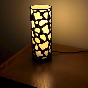 Op de afbeelding: Een zwarte tafellamp met een witte, abstracte print op de kap. De lamp is aan en geeft een warm, wit licht.