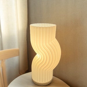 Op de afbeelding: Een witte keramische tafellamp met een golvend, geribbeld ontwerp. De lamp is aan en verlicht de kamer.