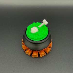 Op de afbeelding: Een kleine zwarte ketel met een groene, getextureerde bovenkant, versierd met een witte schedel en bot. De ketel staat op een oranje, vlamvormige basis, een decoratief item met Halloween-thema.