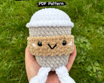 May include: A crocheted coffee cup with a white lid and brown band. The cup has a smiling face with two small pink cheeks. The cup is held in a hand against a green grassy background. The text "Crochet Coffee Cup PDF Pattern" is above the cup. The text "By @hannahhcrochets" is below the cup.