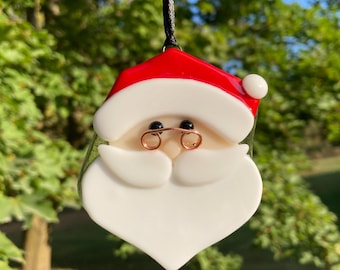Op de afbeelding: Een feestelijke kerstversiering in de vorm van de kerstman. De ornament heeft een rode muts, een witte baard en een bril van koperdraad. Het hangt aan een zwart koord tegen een vage groene achtergrond.