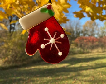 Op de afbeelding: Een rode en witte glazen kerstversiering in de vorm van een want met een sneeuwvlokontwerp. Het ornament heeft een witte manchet met hulst- en bessenaccenten en hangt aan een zwart lint. De achtergrond toont gele bladeren en een blauwe lucht.
