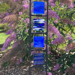 May include: A vertical metal garden stake featuring four square glass panels suspended in a ladder-like frame. The glass pieces display various shades of blue, including deep cobalt, light azure, and textured patterns with white and silver accents.