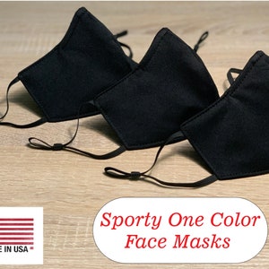 Peut inclure: Trois masques faciaux noirs avec des boucles d'oreille réglables. Les masques sont faits d'un tissu doux et respirant. Le texte "Sporty One Color Face Masks" est imprimé sous les masques. Un petit drapeau américain avec le texte "Made in USA" est dans le coin inférieur gauche de l'image.