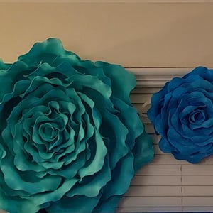 Op de afbeelding: Vier grote papieren bloemdecoraties in roze, blauwgroene en blauwe tinten. De bloemen zijn aan een muur bevestigd, met een jaloezie zichtbaar op de achtergrond. De bloemen vari&euml;ren in grootte, de grootste heeft een diameter van ongeveer 76 cm.