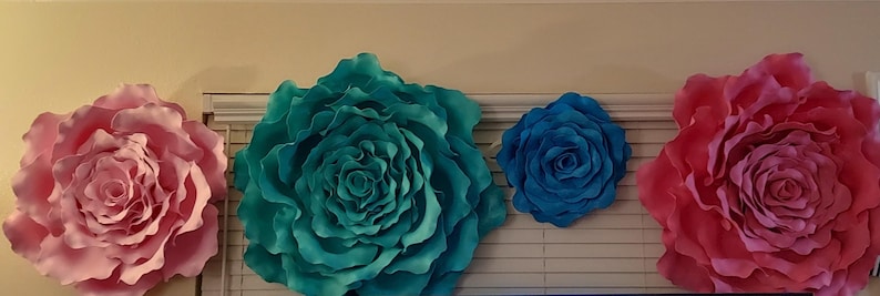 Op de afbeelding: Vier grote papieren bloemdecoraties in roze, blauwgroene en blauwe tinten. De bloemen zijn aan een muur bevestigd, met een jaloezie zichtbaar op de achtergrond. De bloemen vari&euml;ren in grootte, de grootste heeft een diameter van ongeveer 76 cm.