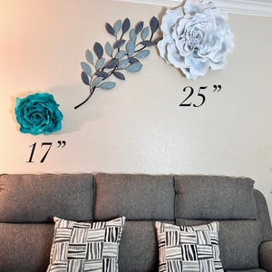 Op de afbeelding: Wanddecoratie met een grote witte en blauwgroene bloem, een blauwe tak met bladeren en twee sierkussens op een grijze bank. De grote bloem is 63,5 cm, en de blauwgroene bloem is 43,2 cm.