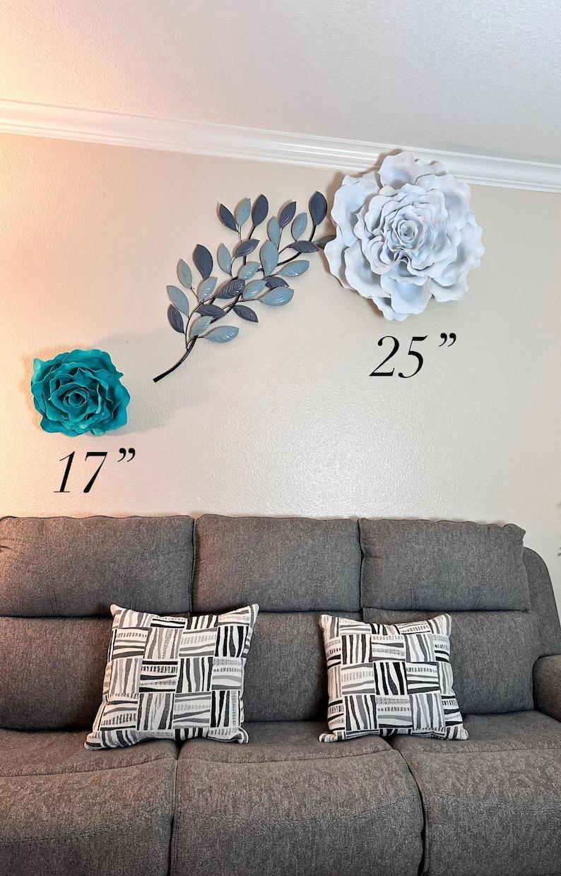 Op de afbeelding: Wanddecoratie met een grote witte en blauwgroene bloem, een blauwe tak met bladeren en twee sierkussens op een grijze bank. De grote bloem is 63,5 cm, en de blauwgroene bloem is 43,2 cm.