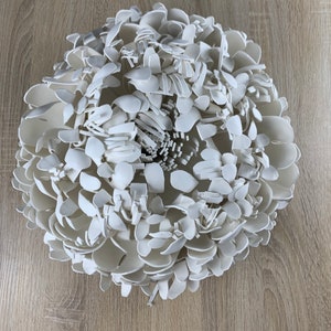 Op de afbeelding: Een grote, witte, driedimensionale bloemensculptuur. De sculptuur is samengesteld uit talrijke bloemblad-vormige stukken, waardoor een gelaagde en getextureerde uitstraling ontstaat. De bloem is gecentreerd en lijkt van keramiek of een vergelijkbaar materiaal te zijn.