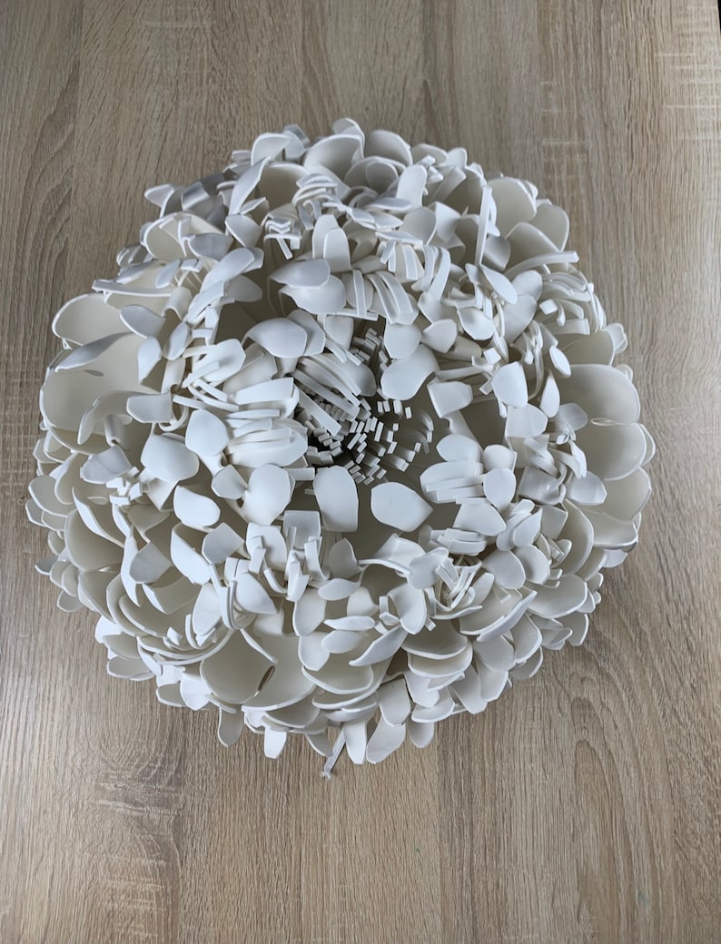 Op de afbeelding: Een grote, witte, driedimensionale bloemensculptuur. De sculptuur is samengesteld uit talrijke bloemblad-vormige stukken, waardoor een gelaagde en getextureerde uitstraling ontstaat. De bloem is gecentreerd en lijkt van keramiek of een vergelijkbaar materiaal te zijn.