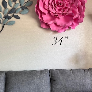 Op de afbeelding: Een grote, levendige roze roos wanddecoratie, ongeveer 86 cm in diameter, is bevestigd aan een neutraal gekleurde muur. Een metalen blad tak in grijstinten en blauw bevindt zich links van de roos. Een grijze bank met patronen kussens staat eronder.