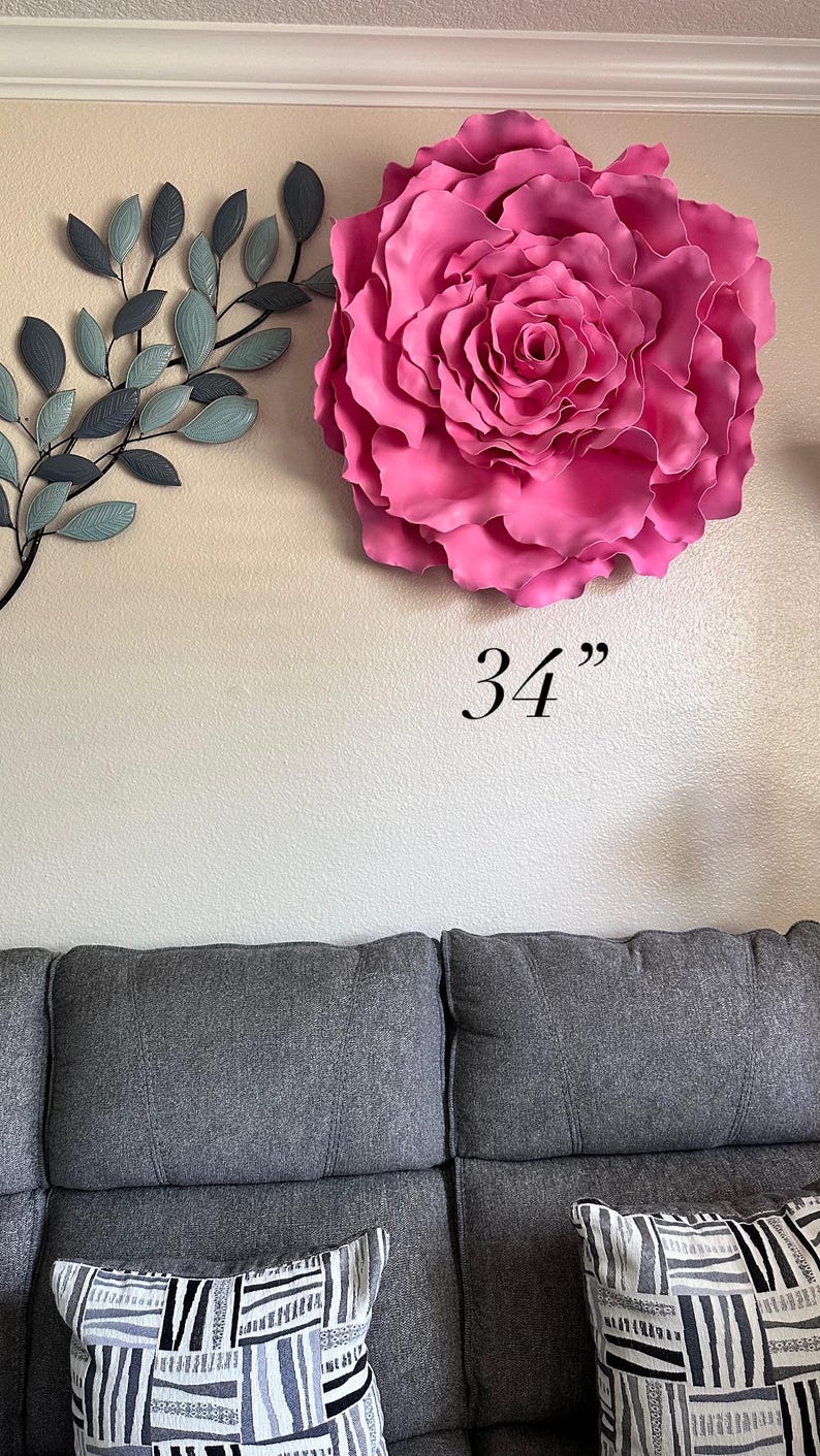 Op de afbeelding: Een grote, levendige roze roos wanddecoratie, ongeveer 86 cm in diameter, is bevestigd aan een neutraal gekleurde muur. Een metalen blad tak in grijstinten en blauw bevindt zich links van de roos. Een grijze bank met patronen kussens staat eronder.