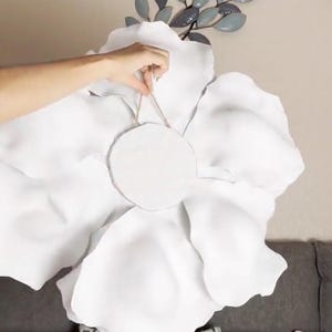 Op de afbeelding: Een grote, witte stoffen bloem voor wanddecoratie, met een cirkelvormig midden. De bloem wordt door een hand vastgehouden, met een neutrale muur en decoratieve metalen bladeren op de achtergrond. De bloemblaadjes zijn zacht en getextureerd.