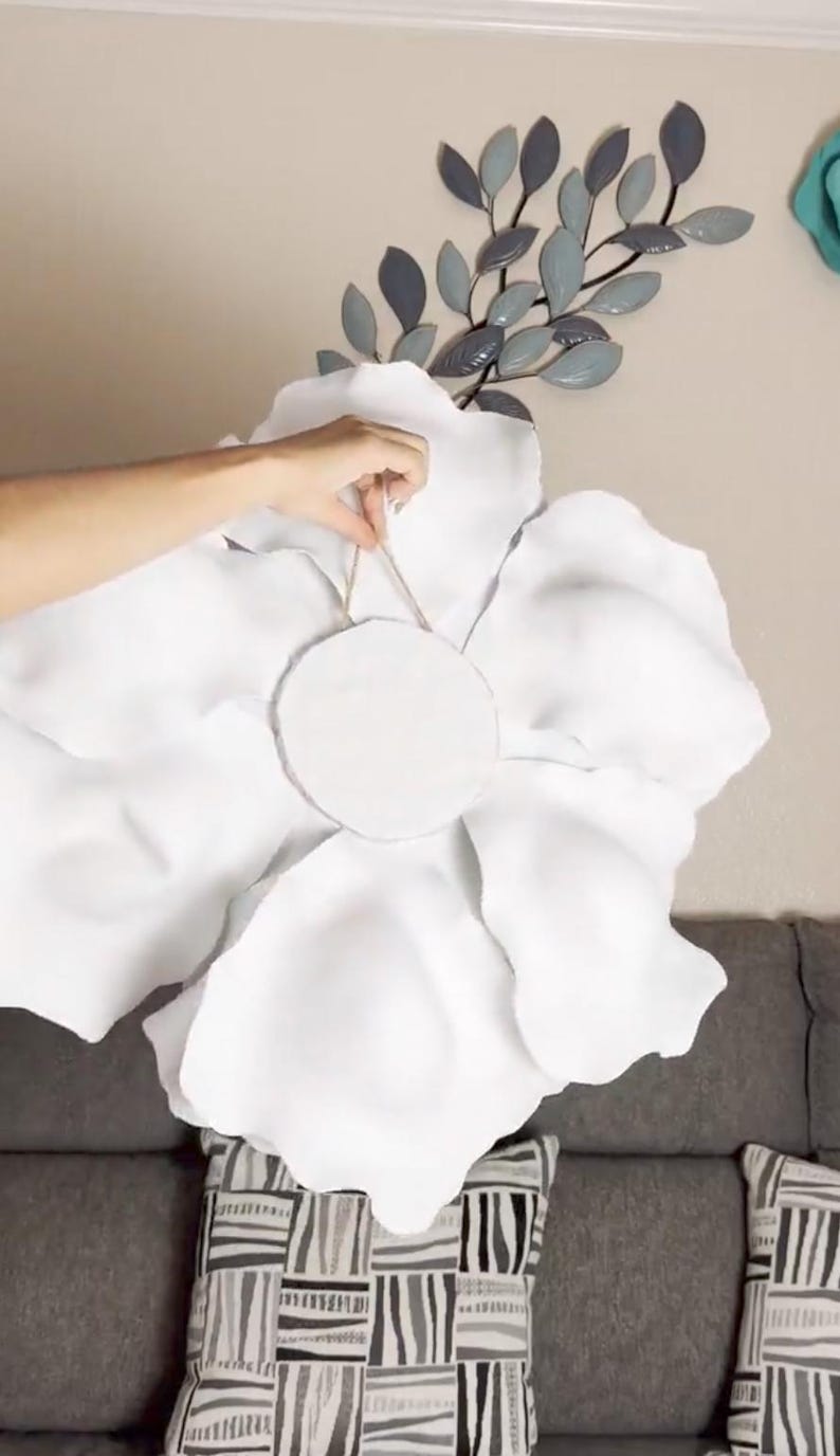 Op de afbeelding: Een grote, witte stoffen bloem voor wanddecoratie, met een cirkelvormig midden. De bloem wordt door een hand vastgehouden, met een neutrale muur en decoratieve metalen bladeren op de achtergrond. De bloemblaadjes zijn zacht en getextureerd.