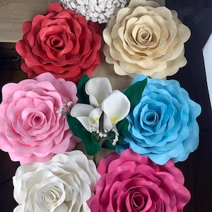 Op de afbeelding: Een assortiment van grote, kleurrijke papieren bloemen, waaronder rode, cr&egrave;me, roze, blauwe, witte en fuchsia rozen. Ook aanwezig zijn witte calla lelies en een witte, getextureerde, cirkelvormige bloemstuk. De bloemen zijn gerangschikt op een houten oppervlak.