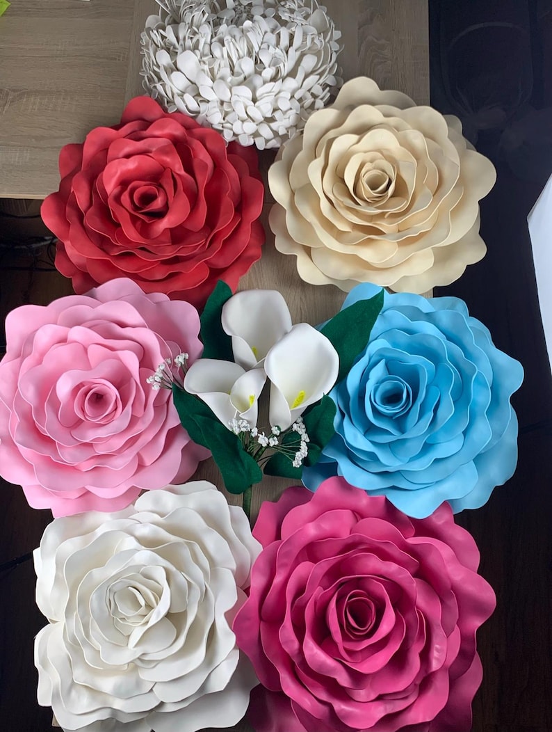 Op de afbeelding: Een assortiment van grote, kleurrijke papieren bloemen, waaronder rode, cr&egrave;me, roze, blauwe, witte en fuchsia rozen. Ook aanwezig zijn witte calla lelies en een witte, getextureerde, cirkelvormige bloemstuk. De bloemen zijn gerangschikt op een houten oppervlak.