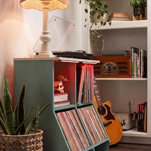 May include: A teal record storage cabinet with angled shelves, filled with vinyl records. A white lamp with a woven shade sits atop the cabinet, next to a record player. A guitar leans against a bookshelf.