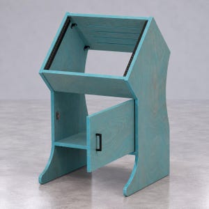 Op de afbeelding: Een turquoise houten studiostandaard met een schuin bovenstuk voorzien van zwarte metalen montagerails en een onderste opbergvak met een enkele deur. De deur is uitgerust met een kleine zwarte handgreep en staat gedeeltelijk open.