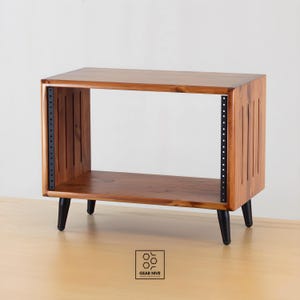 Op de afbeelding: Een houten audio-rack met een natuurlijke afwerking. Het rack heeft een rechthoekige vorm met een plank en een open voorkant. Het heeft zwarte metalen rails voor het monteren van apparatuur en zwarte taps toelopende poten. De zijkanten hebben verticale groeven.