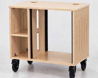 May include: A light-coloured wooden studio rack stand on four black caster wheels. The unit features a flat top surface with two circular cutouts, a side storage section with one shelf, and a central open bay with black metal rack rails.