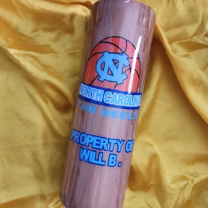 May include: A cylindrical tumbler with a wood grain design. The tumbler features the North Carolina Tar Heels logo and text that reads "NORTH CAROLINA TAR HEELS" and "PROPERTY OF WILL B." in blue.