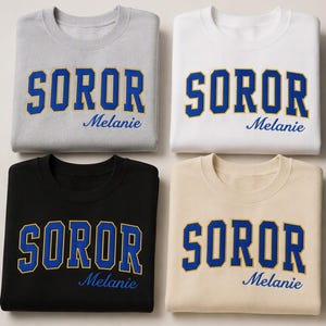 Op de afbeelding: Vier sweaters met ronde hals in grijs, wit, zwart en beige. Elk exemplaar toont het woord SOROR in grote blauwe blokletters met een gele omlijning, en de naam Melanie in blauw cursief schrift onder de hoofdtekst op de voorzijde.