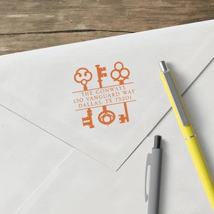 May include: A white envelope with an orange ink stamp featuring key designs and the text "THE CONWAYS 130 VANGUARD WAY DALLAS, TX 75201". Two pens, one yellow and one gray, are on the right side of the envelope.