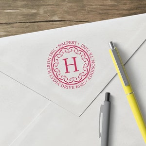 May include: A white envelope with a pink custom address stamp. The stamp is circular with the letter "H" in the center, surrounded by decorative flourishes and the address: 7083 North Lydia Drive #3925 Addison, TX 75001. Two pens are next to the envelope.