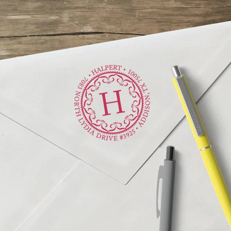 May include: A white envelope with a pink custom address stamp. The stamp is circular with the letter "H" in the center, surrounded by decorative flourishes and the address: 7083 North Lydia Drive #3925 Addison, TX 75001. Two pens are next to the envelope.