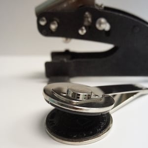 May include: Close-up of a black and silver embossing seal with a circular die. The seal is pressing down on a dark coin-like object. A second, larger seal is in the background.
