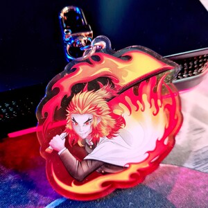 May include: A clear acrylic keychain featuring a character with fiery orange hair and red eyes, holding a red sword. The character is surrounded by a red and orange flame design. The keychain has a silver clasp.