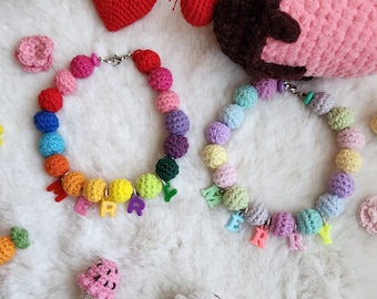 Könnte beinhalten: Zwei bunte gehäkelte Halsketten mit dem Wort "Merry" in bunten Perlen. Die Halsketten liegen auf einer weißen flauschigen Oberfläche mit anderen gehäkelten Gegenständen, darunter ein Herz, eine Karotte und ein Cupcake.