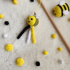Pu&ograve; includere: Un giocattolo a forma di ape fatto a crochet con filati neri e bianchi che pendono dal suo corpo. L'ape &egrave; gialla e nera con un viso bianco. Il giocattolo &egrave; su una superficie bianca e pelosa con altre api fatte a crochet e piccole palline gialle e nere.