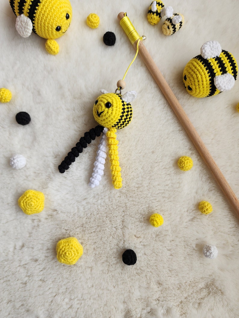 Pu&ograve; includere: Un giocattolo a forma di ape fatto a crochet con filati neri e bianchi che pendono dal suo corpo. L'ape &egrave; gialla e nera con un viso bianco. Il giocattolo &egrave; su una superficie bianca e pelosa con altre api fatte a crochet e piccole palline gialle e nere.