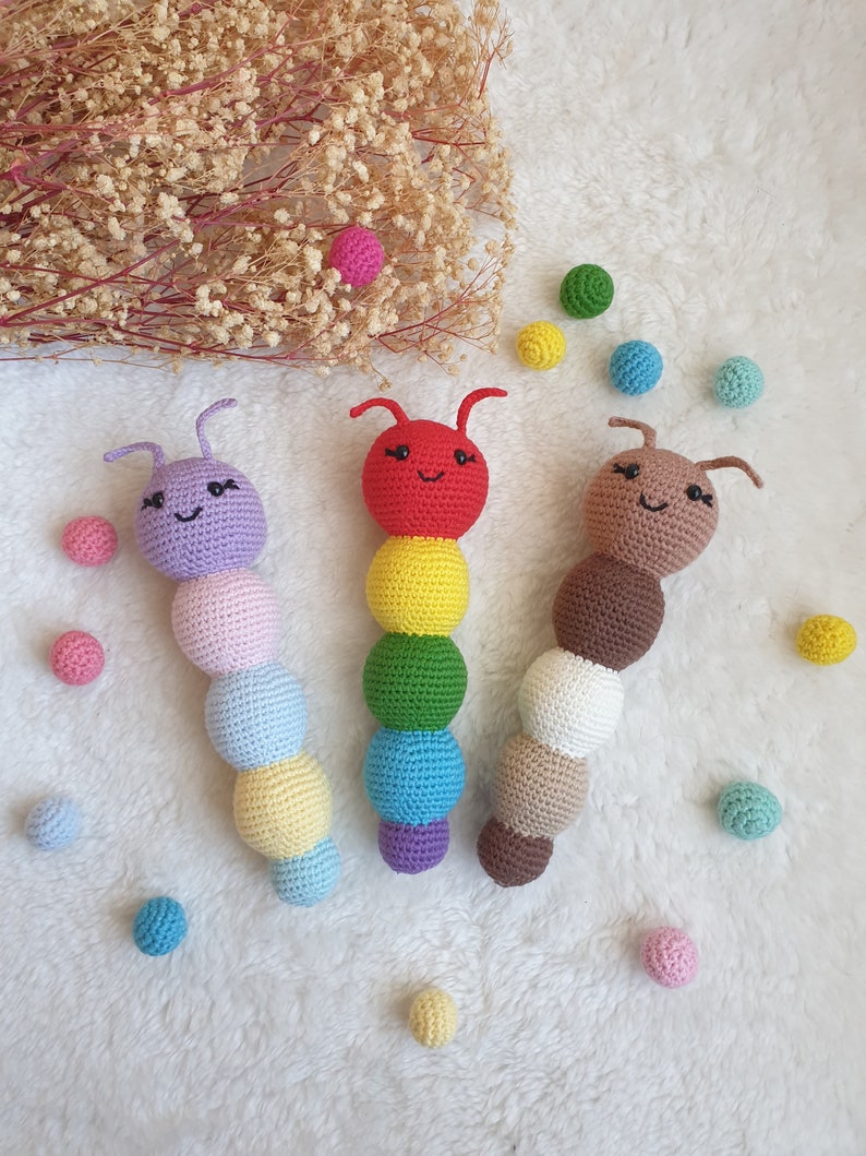 May include: Three crocheted caterpillar toys with colourful bodies and smiling faces. The caterpillars are made of different coloured yarn and have small black eyes. The toys are surrounded by small colourful balls.