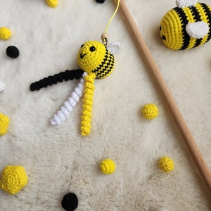 Pu&ograve; includere: Un giocattolo a forma di ape in crochet con strisce nere e gialle appeso a un bastone di legno. L'ape ha un corpo bianco e giallo e occhi neri. Ci sono diverse altre api in crochet e palline gialle e nere sparse intorno al giocattolo a forma di ape.