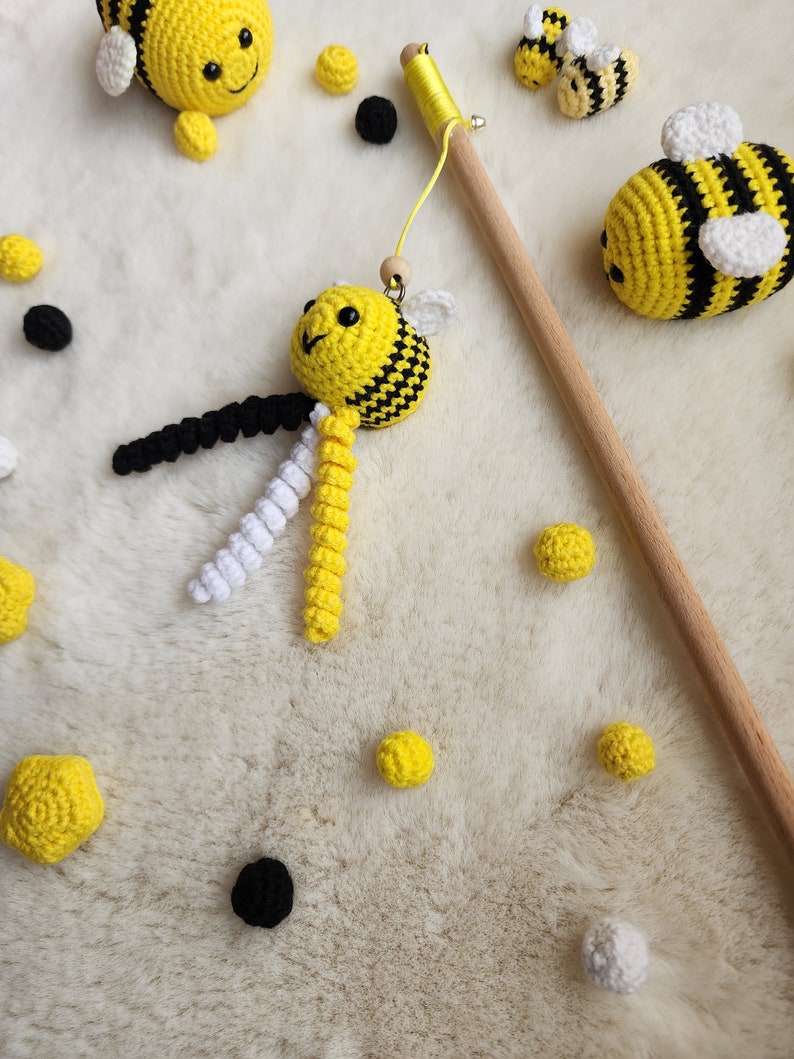 Pu&ograve; includere: Un giocattolo a forma di ape in crochet con strisce nere e gialle appeso a un bastone di legno. L'ape ha un corpo bianco e giallo e occhi neri. Ci sono diverse altre api in crochet e palline gialle e nere sparse intorno al giocattolo a forma di ape.