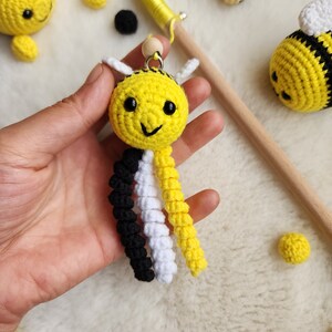 Pu&ograve; includere: Un giocattolo ape all'uncinetto realizzato con filato giallo, nero e bianco. L'ape ha un viso sorridente e una piccola perlina di legno per la testa. L'ape &egrave; appesa a un bastoncino di legno con filato giallo.