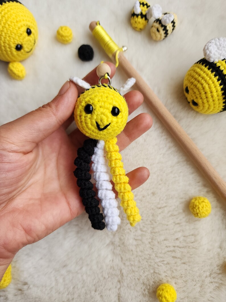 Pu&ograve; includere: Un giocattolo ape all'uncinetto realizzato con filato giallo, nero e bianco. L'ape ha un viso sorridente e una piccola perlina di legno per la testa. L'ape &egrave; appesa a un bastoncino di legno con filato giallo.