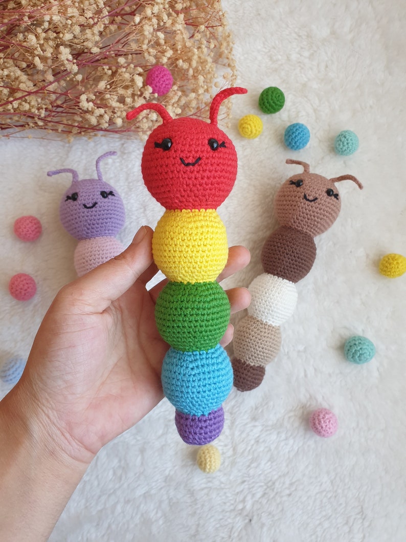 May include: Three crocheted caterpillar toys. The first is red, yellow, green, blue, and purple. The second is purple and the third is brown. Each caterpillar has a smiling face with black eyes and a black mouth.