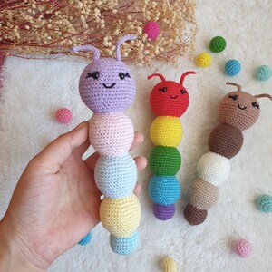 May include: Three crocheted rainbow-coloured caterpillars with smiling faces. The caterpillars are made of different coloured yarn, including purple, pink, blue, yellow, green, red, brown, and white.